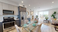 This single-floor residence showcases a contemporary design with a cohesive open-concept layout connecting the kitchen, dining, and living areas. Light wood flooring runs throughout, complementing the bright white walls and recessed lighting that create a clean, airy atmosphere.

The kitchen is well-appointed with two-tone cabinetry — soft grey upper cabinets paired with slate blue lower cabinets — along with white quartz countertops, a full suite of stainless steel appliances including a gas range, built-in microwave, and a French door refrigerator with an ice dispenser. A glass-top dining table sits adjacent to the kitchen, comfortably seating four.

The living room is generously proportioned, featuring recessed lighting and a sliding glass door that opens to a private balcony with open outdoor views. Natural light pours in, enhancing the inviting atmosphere.

Two bedrooms are present; the primary bedroom features large corner windows overlooking mature trees, a mirrored closet, and direct balcony access. The second bedroom is well-lit with its own artwork wall and ample space.

Both bathrooms reflect a minimalist aesthetic, featuring large-format tile flooring, dark countertops, under-mount sinks, backlit mirrors, and a tub-shower combination with glass enclosures.