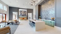 Located at 2950 Richmond Row Drive in Suwanee, Georgia, this contemporary two-story amenity facility showcases a sophisticated design that blends modern aesthetics with welcoming communal spaces. The ground floor greets visitors with a dramatic, double-height lobby featuring soaring ceilings, large-format white tile flooring in a herringbone pattern, and an abundance of natural light streaming through floor-to-ceiling windows. A striking wet bar area with a white quartz island, bar seating, and an iridescent stacked-tile accent wall serves as a central focal point, complemented by sputnik-style chandeliers overhead. A cozy lounge area with a fireplace set against large-format gray tile, a wall-mounted television, and plush seating invites relaxation, while an adjacent sunlit sitting area offers direct views of the outdoor pool. A dedicated fitness center with glass-panel doors is also accessible from the main level. The second floor continues the refined aesthetic with warm wood-look tile flooring, open lounge areas furnished with mid-century modern velvet seating, curated organic wall art, and a built-in beverage station with a wine cooler. Wrought iron railings line the open staircase connecting both levels, reinforcing the space's polished, upscale character throughout.