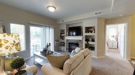 This single-story residence offers a thoughtfully designed layout across several well-defined living areas. The living room serves as a central gathering space, anchored by a stone-surround fireplace framed by built-in display shelving on either side. A wall-mounted television sits above the mantle, while a sliding glass door fills the room with natural light and provides direct access to an outdoor patio. A secondary sitting area features additional sliding door access to the outdoors, extending the sense of openness throughout.

The kitchen is equipped with dark espresso cabinetry, granite-style countertops, a tile backsplash, and a full suite of stainless steel appliances including a gas range, microwave, and refrigerator. Wood-look flooring runs through the kitchen and connects to a dedicated dining area, which accommodates a round table beneath a pendant light fixture. Two bedrooms are present — a primary bedroom with carpet flooring, natural light from large windows, and ample space, alongside a secondary bedroom with carpet and windows overlooking greenery. Two full bathrooms feature wood-look flooring, soaking tubs, and dark vanity cabinetry with stone-style countertops.