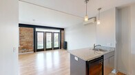 Located at 310 W Sixth St in the heart of Royal Oak, Michigan, this efficiently designed studio seamlessly blends industrial character with modern finishes. The open living area makes a striking first impression with warm hardwood flooring, exposed brick accent walls, and bold navy trim that frames a set of black French doors. These full-length doors invite abundant natural light while offering views of the surrounding neighborhood. Vintage cast-iron radiators on either side of the living space add an authentic industrial charm, while a ceiling fan provides year-round comfort. The kitchen is a functional highlight, centered around a granite-topped island with a stainless steel sink and stylish pendant lighting overhead. White upper cabinetry, stone countertops, and black appliances — including a refrigerator and range — create a clean, contemporary cooking space. A wall-mounted mini-split unit ensures efficient climate control. A separate sleeping area features continuation of the hardwood floors, a ceiling fan, and a sliding-door closet for ample storage. The bathroom is finished with a white vanity, matte black fixtures, and dark tile in the walk-in shower, offering a polished, modern aesthetic. This thoughtfully appointed studio offers an ideal urban living experience in a vibrant Royal Oak location.