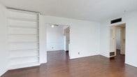 This single-floor space offers a clean, open layout designed for functional everyday living. The main living area features warm hardwood flooring that extends throughout, complementing the bright white walls and textured ceiling. A notable built-in shelving unit lines one wall, providing ample storage and display space while adding a structured architectural element to the room.

An open passageway connects the living area to the kitchen, creating a natural flow between the two spaces. The kitchen is visible through the opening and includes white cabinetry, a ceiling fan for ventilation, and a full-size refrigerator, all finished in a consistent white palette that keeps the space feeling light and cohesive.

The textured popcorn ceiling runs throughout both areas, a characteristic period detail. The white walls throughout provide a neutral backdrop adaptable to a variety of furnishing styles. The overall ambiance is minimal and bright, with the open-plan configuration and consistent flooring lending a sense of continuity and spaciousness across the interior.