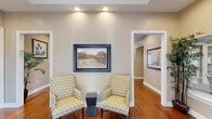 Located at 616 Highway 90 in Waveland, Mississippi, this single-floor community space offers a warm and inviting atmosphere defined by thoughtful architectural details and high-quality finishes. Rich hardwood flooring flows seamlessly throughout the entire space, complementing the neutral taupe wall tones and crisp white trim that frame each room.

The standout architectural feature is the striking exposed wood beam ceiling, which adds rustic elegance and visual depth. Recessed lighting and ceiling fans are integrated throughout, ensuring the space is both well-lit and comfortable year-round.

The two living areas are generously proportioned and thoughtfully arranged. The main living area serves as the focal point of the space, furnished with a plush sectional sofa, accent chairs, and an area rug anchoring the seating arrangement. A dark wood entertainment unit with built-in shelving provides both functional storage and decorative display. French doors allow natural light to filter in and offer access to adjacent areas.

A secondary seating area provides a more intimate setting, featuring a pair of patterned accent chairs, a small side table, and framed wall art. Additional connector hallways lead to transitional nooks, each furnished with accent consoles and artwork, contributing to a cohesive and polished overall design. The space exudes a refined yet welcoming character throughout.