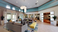 This single-floor community amenity space is designed with a modern, resort-inspired aesthetic that balances comfort and functionality. The centerpiece is a generous open lounge area crowned by soaring double-height ceilings accented with bold teal crown molding and warm neutral walls. Oversized globe pendant light fixtures hang overhead, casting a soft, welcoming glow across the seating area below, which is furnished with contemporary gray sofas, teal accent chairs, and a vibrant geometric area rug in shades of blue, teal, and yellow. Light-toned wood-look tile flooring runs throughout, adding warmth and cohesion to the open layout. Floor-to-ceiling glass sliding doors line one wall, allowing abundant natural light to flood the interior while offering direct access to a sunlit outdoor patio. Sheer linen drapes frame the expansive windows, softening the transition between indoors and out. Tall white planters with striking architectural plants are positioned throughout, enhancing the fresh, tropical ambiance. Adjacent to the main lounge, a multipurpose room is accessible through elegant frosted-glass panel doors and features hardwood flooring, track lighting, a wall-mounted television, and flexible furnishings. A fitness center with mirrored walls and cardio equipment is also visible from the lounge, rounding out this well-appointed communal space.