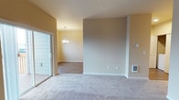 This single-floor residence offers a clean, move-in-ready layout designed with comfort and functionality in mind. The living area is finished with plush wall-to-wall carpeting in a neutral beige tone, creating a warm and inviting atmosphere. A large sliding glass door opens directly to an outdoor deck, flooding the space with natural light and providing convenient indoor-outdoor access.

Adjacent to the main living area, a dedicated dining space is distinguished by rich hardwood-style flooring and anchored by an elegant chandelier overhead, adding a refined touch to everyday gatherings. The transition between the two areas is defined by a subtly raised ceiling line and a structural accent wall finished in a soft taupe hue, lending the space a sense of architectural dimension.

Warm yellow-toned walls throughout complement the neutral palette and contribute to a bright, cheerful ambiance. The space is equipped with a wall-mounted heater, ensuring year-round comfort. Clean white baseboards and trim provide crisp contrast against the warmer wall tones, completing the polished, cohesive interior aesthetic. Overall, this single-floor layout balances practical design with comfortable living in a well-lit, thoughtfully finished environment.