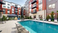 Located at 2031 Maywill St in Richmond, Virginia, this contemporary residential community showcases a thoughtfully designed outdoor courtyard that serves as the centerpiece of the property. The multi-story building features a striking exterior combining bold red and charcoal gray siding with light neutral accents, creating a modern, urban aesthetic complemented by private balconies on upper floors.

The courtyard is anchored by a sparkling swimming pool surrounded by a generous pool deck furnished with lounge chairs, offering residents a resort-like retreat. Adjacent to the pool, a spacious patio area provides outdoor dining with white tables and chairs arranged for communal gatherings, making it ideal for relaxing or entertaining.

A dedicated recreational area features a cornhole board set on wide-format concrete pavers, framed by raised stone-clad planter walls filled with lush ornamental grasses and evergreen shrubs. Landscaped garden beds with a variety of green plantings and decorative gravel line the walkways, adding natural texture and color throughout the courtyard.

A welcoming glass-door entrance, accented by string lights overhead, connects the outdoor space to the building's interior. Covered walkways, black metal railings, and a metal-roofed outbuilding contribute to the cohesive, well-appointed design. The entire outdoor environment reflects a polished, amenity-rich community built for modern urban living in Richmond.