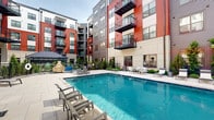 Located at 2031 Maywill St in Richmond, Virginia, this contemporary residential community showcases a thoughtfully designed outdoor courtyard that serves as the centerpiece of the property. The multi-story building features a striking exterior combining bold red and charcoal gray siding with light neutral accents, creating a modern, urban aesthetic complemented by private balconies on upper floors.

The courtyard is anchored by a sparkling swimming pool surrounded by a generous pool deck furnished with lounge chairs, offering residents a resort-like retreat. Adjacent to the pool, a spacious patio area provides outdoor dining with white tables and chairs arranged for communal gatherings, making it ideal for relaxing or entertaining.

A dedicated recreational area features a cornhole board set on wide-format concrete pavers, framed by raised stone-clad planter walls filled with lush ornamental grasses and evergreen shrubs. Landscaped garden beds with a variety of green plantings and decorative gravel line the walkways, adding natural texture and color throughout the courtyard.

A welcoming glass-door entrance, accented by string lights overhead, connects the outdoor space to the building's interior. Covered walkways, black metal railings, and a metal-roofed outbuilding contribute to the cohesive, well-appointed design. The entire outdoor environment reflects a polished, amenity-rich community built for modern urban living in Richmond.