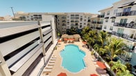This residential community is defined by its striking resort-style outdoor amenity space, thoughtfully designed to maximize both comfort and visual appeal. At the heart of the complex lies a generously sized, decoratively shaped swimming pool with crystal-clear water, serving as the centerpiece of a well-appointed courtyard. The pool deck is lined with numerous lounge chairs and sun loungers, offering ample space for relaxation under open skies. Vibrant red-orange shade umbrellas and covered cabana structures provide welcome relief from the sun, creating inviting spots for leisure throughout the day. Lush tropical palm trees frame the entire space, lending a warm, resort-like atmosphere that evokes a sense of escape. The surrounding multi-story building features a modern architectural design with a neutral facade, tiled rooflines, and private balconies that overlook the courtyard below. An integrated parking structure is also visible within the complex, reflecting practical and well-planned design. The community's overall ambiance is bright, open, and welcoming, with the combination of contemporary architecture, tropical landscaping, and premium outdoor amenities creating a cohesive and attractive living environment.