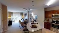 This single-floor residence offers a thoughtfully designed open-concept layout that seamlessly connects its primary living spaces. The kitchen serves as a central hub, featuring granite countertops on a curved breakfast bar with bar seating, rich dark cabinetry, and pendant lighting that adds a refined touch. Track lighting overhead provides additional illumination throughout the main living area.

The open living and dining zone flows naturally from the kitchen, with a carpeted dining area accommodating a round table and upholstered seating. The main living room receives generous natural light through large sliding glass doors that lead to a private balcony, offering outdoor views and bringing a bright, airy quality to the space. Warm, neutral tones and soft carpeting contribute to an inviting ambiance throughout.

A secondary sitting or media room is positioned adjacent to the main living area, providing a versatile space for entertainment or relaxation. The layout also includes at least one private bedroom, comfortably furnished and separated from the main living areas for added privacy. A bathroom and closet space are visible within the floor plan, contributing to the overall functionality of this well-organized residence.