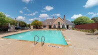 Located at 125 S Murdeaux Ln in Dallas, TX, this single-story community clubhouse presents an inviting and well-appointed gathering space for residents. The building's exterior showcases a charming combination of stone masonry and stucco finishes, topped with a pitched roof featuring a distinctive turret-style accent and chimney, lending the structure a classic, residential character.

Entry to the clubhouse is made through elegant black French doors with multi-pane glass panels, allowing natural light to filter into the interior. A secondary glass-panel door and adjacent window provide additional access points and visibility into the space within.

The centerpiece of the outdoor area is a generously sized swimming pool with clear turquoise water, surrounded by a wide concrete pool deck ideal for lounging. The pool area is enclosed by a black wrought iron security fence with a gated entry, ensuring safety and controlled access. Beyond the pool, mature trees line the perimeter, providing natural shade and a lush green backdrop. A playground area is also visible within the fenced grounds, adding to the community-oriented nature of the property.

The interior entry area features bare concrete flooring and open doorways, suggesting a functional, adaptable layout suitable for community events and gatherings. Overall, the space combines practical amenities with classic architectural appeal in a well-maintained outdoor setting.