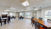 This single-floor community space in Concord, CA (94521) is designed to serve as a shared gathering area. The main room is generously sized, finished with light-colored ceramic tile flooring and a textured ceiling fitted with recessed fluorescent light panels that ensure even illumination throughout. Multiple wooden dining tables with black slatted chairs are arranged across the open floor plan, accommodating groups of various sizes for meals or meetings. A notable feature is a classic upright piano positioned near the windows, adding a social and recreational element to the space. Large sliding and fixed windows line multiple walls, flooding the interior with natural light and offering clear views of the surrounding landscaped grounds and adjacent buildings. A dedicated kitchen area is accessible via a pass-through counter, equipped with light wood cabinetry, a stainless steel range, microwave, refrigerator, and sink — well-suited for catering or communal meal preparation. A secondary, more flexible open area with tile flooring and minimal furnishings provides additional utility space. The overall ambiance is clean, functional, and well-lit, designed to support a variety of community activities in a comfortable, accessible single-level setting.
