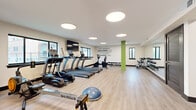 This fitness center offers a thoughtfully designed exercise space spread across a single open floor. The room features light wood-style flooring, white walls, and a clean, modern aesthetic complemented by circular flush-mount ceiling lights that provide consistent, even illumination throughout. Large black-framed windows line the perimeter, allowing ample natural light and outdoor views to create an energizing workout environment.

The cardio zone is well-appointed with multiple TRUE-brand treadmills, elliptical machines, a recumbent bike, and a Shi-Row rowing machine, all positioned along the windowed wall beneath a wall-mounted flat-screen television. A BOSU balance trainer and additional functional fitness tools occupy the open floor space at the center of the room.

A dedicated strength training area features a functional cable machine, a rack of colorful dumbbells ranging across various weights, a weight bench, and a full-length mirror that visually expands the space. A storage rack stocked with medicine balls, resistance bands, and exercise balls adds further versatility for functional and group-style training. Accent columns in green and blue add pops of color, while a bold motivational wall decal — "If It Doesn't Challenge You, It Doesn't Change You" — reinforces the space's energetic atmosphere. Sliding glass doors provide access to an adjacent outdoor area.
