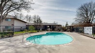 The Trees at Madison, located at 5101 Hackberry Lane in Sacramento, California, offers a well-appointed outdoor community pool area designed for relaxation and recreation. The centerpiece of this amenity space is a generously sized freeform swimming pool with clear, turquoise water, accessible via built-in entry steps and a pool ladder. The surrounding concrete patio deck provides ample room for lounging, with seating and tables arranged for comfort and socializing.

A dedicated outdoor grilling station is conveniently positioned along the pool deck, making the area well-suited for gatherings. The space is fully enclosed by a sturdy wrought iron fence with a gated entry, ensuring both safety and security for residents. Posted pool rules and emergency information signage reflect a commitment to resident well-being.

Mature, towering trees frame the entire area, lending a natural, shaded ambiance that enhances the outdoor experience, particularly during Sacramento's warm seasons. The surrounding landscaping includes manicured green lawns and garden beds that contribute to a serene atmosphere. Single-story residential buildings are visible in the background, integrating the pool area cohesively within the broader community. The pool operates with a posted capacity and set hours, underscoring its role as a structured yet inviting shared amenity within The Trees at Madison community.