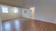 This single-floor space is designed with a clean, functional layout that prioritizes open living. The main living area is immediately welcoming, showcasing gleaming hardwood floors in a warm honey tone that extend throughout the space, reflecting natural and artificial light to create a bright, airy atmosphere. Crisp white trim and neutral beige walls provide a versatile backdrop suitable for a variety of décor styles.

The open floor plan allows for a natural flow between the living area and the adjoining spaces. A pass-through window connects the living area to the kitchen, which features light wood cabinetry and includes a full-size refrigerator, offering a practical and efficient workspace. An additional room visible through a doorway provides further utility, with its own lighting and what appears to be additional storage or utility space.

Ceiling-mounted HVAC vents and floor-level registers are strategically placed throughout, ensuring consistent climate control across the entire floor. A window in the living area allows natural light to filter in, further brightening the interior. The straightforward, well-maintained design of this space makes it a practical and comfortable environment, ready for immediate use.