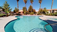 This single-level outdoor amenity space offers a well-appointed resort-style retreat designed for relaxation and leisure. The centerpiece is a generously sized freeform swimming pool with striking blue tile accents and crystal-clear water, accompanied by an adjacent circular spa that provides a dedicated area for hydrotherapy and unwinding. The smooth concrete pool deck surrounds the water on all sides, offering ample space for lounging and movement. A covered patio area with stone-accented columns and a pergola-style roof provides shaded outdoor dining, complete with a glass-top table and chairs — an ideal spot to enjoy the outdoors while staying cool. The landscaping features a combination of tall fan palms, desert shrubs, and manicured greenery that lend a lush, tropical character to the desert surroundings. The fenced perimeter ensures privacy and safety while maintaining open sightlines to the surrounding community architecture, characterized by warm stucco exteriors and clay tile rooflines. Natural light floods the entire space throughout the day, and the open-sky setting enhances the bright, airy ambiance. Overall, the area conveys a welcoming, community-oriented atmosphere with a strong emphasis on outdoor living in a sun-drenched desert environment.