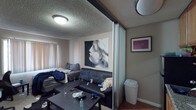 This single-floor unit offers a functional layout designed to maximize its compact footprint. The living area serves as the central space, featuring hardwood-style flooring, a textured popcorn ceiling, and a window fitted with vertical blinds that allows natural light to filter in. A frosted glass sliding partition divides the living area from the kitchen, creating a flexible sense of separation while maintaining an open feel between the two spaces.

The kitchen is efficiently arranged with warm-toned wood cabinetry providing ample overhead and base storage. Stainless steel appliances include a gas range with oven and an over-the-range microwave, complemented by a full-size refrigerator and a granite-style countertop with a sink. A hallway off the kitchen leads to additional storage space, including what appears to be a walk-in closet area, along with open wall shelving.

The bathroom features white tile walls accented by a decorative horizontal band of multicolored mosaic tiles. A bathtub and shower combination is enclosed by a sliding frosted glass door with chrome framing, accompanied by a standard toilet. Overall, the unit presents a utilitarian design with practical storage solutions throughout.