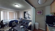 This single-floor unit offers a functional layout designed to maximize its compact footprint. The living area serves as the central space, featuring hardwood-style flooring, a textured popcorn ceiling, and a window fitted with vertical blinds that allows natural light to filter in. A frosted glass sliding partition divides the living area from the kitchen, creating a flexible sense of separation while maintaining an open feel between the two spaces.

The kitchen is efficiently arranged with warm-toned wood cabinetry providing ample overhead and base storage. Stainless steel appliances include a gas range with oven and an over-the-range microwave, complemented by a full-size refrigerator and a granite-style countertop with a sink. A hallway off the kitchen leads to additional storage space, including what appears to be a walk-in closet area, along with open wall shelving.

The bathroom features white tile walls accented by a decorative horizontal band of multicolored mosaic tiles. A bathtub and shower combination is enclosed by a sliding frosted glass door with chrome framing, accompanied by a standard toilet. Overall, the unit presents a utilitarian design with practical storage solutions throughout.