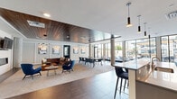 Located at 3250 W 66th St in Edina, Minnesota, this single-floor community space offers a sophisticated, open-concept layout that seamlessly blends a fully equipped kitchen with a welcoming living area. The kitchen is a standout feature, boasting sleek gray cabinetry, a large center island with bar seating, stainless steel appliances including a professional-grade gas range with a stainless hood, and a striking white textured tile backsplash. Pendant lighting in matte black adds a modern contrast to the bright, white surfaces. Large floor-to-ceiling windows flood the space with natural light and provide views of an outdoor patio area, enhancing the open, airy feel. Large-format tile flooring transitions into warm wood-tone flooring throughout the living area. The lounge features a rich wood-paneled ceiling that adds warmth and architectural character, complemented by a wall-mounted fireplace, a flat-screen TV, and curated artwork. Comfortable seating—including a leather sectional and bold blue accent chairs—creates an inviting gathering atmosphere. Structural columns and an open floor plan contribute to the spacious, loft-like character of this well-appointed community amenity space.