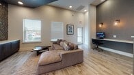 This well-appointed single-floor amenity space combines modern design with practical functionality across several distinct areas. The lounge area anchors the space with plush, oversized seating arranged around a live-edge coffee table, complemented by light wood-look tile flooring and soaring ceilings that create an open, airy atmosphere. A striking accent wall featuring a metallic mosaic tile backsplash frames a wall-mounted display unit with dark cabinetry, serving as a bold focal point in the lounge. Natural light flows generously through multiple large windows fitted with blinds, keeping the interior bright and connected to the outdoors. The business center is set apart by its moody, textured dark accent walls and warm Edison-style pendant lighting, housing multiple computer workstations ideal for productive work sessions. A tall counter with bar-height seating provides an additional workspace option. Adjacent to the main lounge, the game room features a full-size pool table beneath a sleek rectangular pendant light, with large bay windows that flood the area with natural light. Framed artwork and mirrors decorate the walls throughout, adding a polished, curated feel. Together, these areas form a cohesive, stylish community space designed to accommodate both relaxation and productivity.