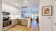 This efficiently designed 2-bedroom, 1-bathroom residence is laid out on a single floor, maximizing every square foot through a thoughtful open-concept plan. Light wood-tone flooring runs throughout the main living areas, creating a warm, cohesive aesthetic that connects the kitchen, dining, and living spaces.

The kitchen is a standout feature, outfitted with dual-tone cabinetry—white upper cabinets paired with light wood lower cabinets—complemented by a white subway tile backsplash and white quartz countertops. A central peninsula with an undermount sink and stainless steel appliances, including a refrigerator, range, microwave, and dishwasher, provides both functionality and style. Striking matte black pendant lights hang above the peninsula, adding a contemporary design element.

The living area benefits from abundant natural light streaming through large French doors that open to a private outdoor area, with a ceiling fan overhead enhancing comfort. Both bedrooms are carpeted and well-lit by sizeable windows with blinds. The primary bedroom comfortably accommodates a queen bed and includes a ceiling fan. The full bathroom features white cabinetry, a quartz vanity, a large mirror, and tile flooring. A dedicated closet and additional storage complete the space.