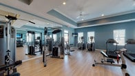 Located at 75 Siskin Cir in Hampstead, NC, this community fitness center offers a comprehensive, well-equipped space designed to support a wide range of workout routines. The single-floor facility features a spacious main exercise area finished with light hardwood-style flooring, tray ceilings with recessed lighting, and ceiling fans that maintain a comfortable environment throughout. The walls are painted in a calming steel blue tone, lending the space a clean and modern aesthetic.

The cardio zone is equipped with multiple treadmills and elliptical trainers, each positioned near windows fitted with white blinds that allow natural light to filter in. Wall-mounted televisions are placed throughout the room to keep members entertained during workouts. The strength training area includes a cable machine system and various resistance equipment, complemented by large floor-to-ceiling mirrors that visually expand the space and allow for proper form checks.

A dedicated free weights section houses a full rack of dumbbells, kettlebells in graduated sizes, and stability balls and BOSU trainers for functional fitness. A flat weight bench rounds out the strength offerings. Adjacent to the main gym floor, a separate children's room provides a welcoming area for young ones, furnished with colorful rounded seating, a chalkboard easel, a large flat-screen TV, and a vibrant area rug.