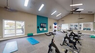 This single-floor fitness studio is thoughtfully designed to support a wide range of exercise activities in a bright, open environment. The space is defined by its striking vaulted ceilings, which lend an airy sense of volume, and are complemented by multiple ceiling fans and recessed linear lighting that ensure consistent illumination throughout. A bold teal accent wall serves as the room's focal point, anchoring a wall-mounted flat-screen television alongside organized storage for workout accessories such as resistance bands and printed materials. Inspirational phrases, including "inhale, exhale" and "NAMA'Slay," are displayed on the walls, setting an encouraging and motivational tone. A row of stationary spin bikes is arranged across the carpeted floor, providing ample space for group cycling sessions. Along one wall, floor-to-ceiling mirrors visually expand the room and allow exercisers to monitor their form. A dedicated rack holds an assortment of stability and medicine balls in various sizes, while exercise mats in vibrant blue are available for stretching and floor work. Large framed windows line multiple walls, allowing natural light to filter in and offering views of the surrounding outdoor environment. The overall ambiance is clean, energetic, and welcoming, making it a versatile space well-suited for both group fitness classes and individual workouts.