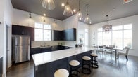 This contemporary coffee lounge is thoughtfully designed to balance functionality with modern aesthetics. The space is anchored by a striking, elongated white quartz bar counter that contrasts boldly against dark cabinetry, offering ample seating with upholstered round barstools. Above, a curated arrangement of clear glass pendant lights and warm recessed lighting hangs from elevated ceilings, creating an inviting, well-lit atmosphere throughout the day. The kitchen area features dark flat-front cabinetry, a stainless steel sink, and a classic white subway tile backsplash, lending the space a clean and polished look. Large windows along the perimeter flood the interior with natural light, enhancing the open and airy feel of the lounge. Polished concrete floors add an industrial edge that complements the overall modern design language. A dining area with metal-framed chairs and tables sits adjacent to the bar, providing a comfortable setting for small gatherings or individual use. The cohesive blend of dark and light tones, high ceilings, and carefully selected fixtures makes this coffee lounge a sophisticated and welcoming communal space.