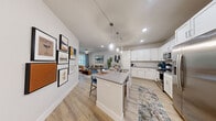 This thoughtfully designed single-floor residence offers a seamless blend of modern style and functional living. The open-concept layout connects the kitchen, dining, and living areas into one cohesive space, ideal for both everyday living and entertaining. The kitchen is a standout feature, boasting crisp white shaker cabinets, grey quartz countertops, a classic subway tile backsplash, and a full suite of stainless steel appliances including a French door refrigerator, electric range, built-in microwave, and dishwasher. A center island with pendant lighting and bar seating serves as both a prep space and casual dining hub. Light wood-look flooring flows throughout the main living areas, complementing the neutral, light-filled palette. The living room is bright and welcoming, anchored by large windows with natural light and a ceiling fan overhead. The bedroom offers plush carpeting, ample closet space, and a serene atmosphere with natural light filtering through a window. The bathroom is finished with grey quartz countertops, a white vanity, a large framed mirror, and a shower-tub combination. Clean lines, recessed lighting, and a cohesive neutral color scheme give the entire space a polished, contemporary feel.