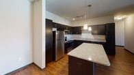 This single-floor residence at 17571–17587 Dodd Blvd in Lakeville, Minnesota offers a well-organized layout designed for comfortable everyday living. The open-concept living and kitchen area serves as the centerpiece of the unit, where rich hardwood-style flooring extends throughout and natural light streams in through multiple windows, casting warm patterns across the floor. Ceiling fans are present in the living areas, contributing to both comfort and airflow.

The kitchen is thoughtfully appointed with dark espresso cabinetry reaching toward the ceiling, a full suite of stainless steel appliances including a refrigerator, range, and microwave, and granite countertops that continue onto a substantial kitchen island. Pendant lighting above the island adds a polished, contemporary touch, while track lighting illuminates the workspace behind it.

The bedrooms feature plush carpeting and ceiling fans, with windows fitted with blinds that allow natural light while maintaining privacy. The bathrooms are finished consistently with granite-topped dark vanities, tub-shower combinations with clean white surrounds, and wood-look flooring. The overall aesthetic is modern and cohesive, with neutral tones and quality finishes carried throughout the entire space.