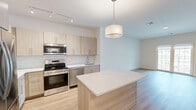 Located at 4180 Sunrise Hwy in Oakdale, NY, this well-appointed one-bedroom, one-bathroom residence offers a thoughtfully designed layout spread across a single living floor. The open-concept kitchen and living area serves as the heart of the home, featuring light wood-grain cabinetry, a crisp white subway tile backsplash, and a full suite of stainless steel appliances including a French door refrigerator, range, microwave, and dishwasher. A large central island with a quartz countertop provides additional prep space and doubles as a casual dining area, illuminated by a modern drum pendant light. Track lighting adds further brightness to the kitchen zone, while light hardwood flooring flows seamlessly throughout the living spaces, enhancing the clean, contemporary aesthetic. The bedroom is generously proportioned and filled with natural light through a wide window that overlooks the building's exterior. Neutral tones and light flooring keep the room feeling airy and versatile. The bathroom is finished with accessibility-conscious grab bars and a clean, minimalist design. The residence is part of a multi-story building complex featuring an expansive landscaped courtyard with patio seating areas, pergolas, and manicured greenery — ideal for outdoor relaxation.