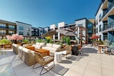 Located at 3100 Lake Villa Dr in Metairie, Louisiana, the Latitude Rooftop Lounge is an expansive open-air terrace designed for sophisticated outdoor living and entertaining. The space is defined by its large-format porcelain tile deck that stretches across multiple distinct seating zones, each thoughtfully curated with premium furnishings including plush white sofas, woven rattan lounge chairs, and warm wood-toned accent tables. Oversized cantilever umbrellas provide shaded retreats throughout the terrace, while a striking wood-framed pergola anchors one corner of the space, adding architectural depth and character. A living green wall adorned with trailing vines serves as a dramatic backdrop, prominently featuring the Latitude Rooftop Lounge signage. Lush potted plants, ornamental trees in soft pink and yellow, and tropical greenery are strategically placed throughout, creating a vibrant garden-like atmosphere under the open Louisiana sky. A dedicated dining area with bistro-style tables and chairs offers a more intimate setting, complemented by string lights that enhance the ambiance during evening hours. Sun loungers positioned along one edge of the terrace invite relaxation, while the surrounding multi-story residential building provides a sense of enclosure and privacy. The overall ambiance is modern, resort-inspired, and ideal for both casual gatherings and elevated social events.