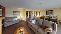 This single-floor community room is designed to serve as a versatile, inviting gathering space. The layout is thoughtfully organized into distinct functional areas that flow naturally into one another.

The main lounge area features warm, neutral-toned carpeting and yellow-painted walls that create a bright, welcoming atmosphere. A standout focal point is the wood-framed electric fireplace set against a bold deep-red accent wall, accompanied by a wall-mounted television above the mantle. Comfortable upholstered seating, including sofas and armchairs, is arranged throughout to encourage relaxed conversation.

A dining area with a wooden table and classic Windsor-style chairs offers space for group meals or activities. The room connects through an arched doorway to additional seating, providing flexibility for various uses.

The kitchen area is functional and well-equipped, featuring light oak cabinetry, a double stainless steel sink, a coil-burner electric range, a microwave, and ample counter space. A checkered tile floor adds a distinct visual element to this zone.

Natural light fills the space through multiple windows dressed with valances, and a set of double glass doors opens directly to an outdoor area, further enhancing the open, community-oriented character of the room.