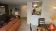 This single-floor residence offers a practical and cohesive layout designed for comfortable everyday living. The living area is carpeted and furnished with ample seating, and is positioned adjacent to a dedicated dining space with a classic wooden table and chairs — ideal for both casual meals and entertaining. A pass-through window connects the living and dining area to the kitchen, creating an open feel that promotes easy interaction between spaces. The kitchen features white cabinetry, overhead lighting, and a functional countertop workspace, keeping the cooking area bright and accessible. A decorative plant placed at the pass-through adds a touch of natural warmth to the interior. The entryway, visible from the living area, features a solid front door that provides a clear sense of arrival and separation between the interior and exterior. A bedroom area with a sliding mirrored closet door offers practical storage with built-in shelving and hanging space. Neutral wall tones and consistent carpeting throughout the main living spaces contribute to a unified and inviting ambiance. The interior lighting is warm and well-distributed, enhancing the overall comfort of the space.