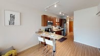 This thoughtfully designed 2-bedroom, 2-bathroom model unit offers a well-organized single-floor layout that balances modern style with everyday functionality. The open-concept kitchen is a standout feature, showcasing rich wood cabinetry, granite countertops, stainless steel appliances, and a breakfast bar with seating — all illuminated by track lighting overhead. Light-toned hardwood-style flooring flows throughout the main living areas, enhancing the bright and airy atmosphere. The living area benefits from generous natural light streaming through large sliding glass doors fitted with vertical blinds, creating a seamless connection to the outdoors. Both bedrooms feature an industrial-chic aesthetic with exposed ductwork overhead, ceiling fans, and large windows that invite natural light. Curated gallery walls add a creative touch to each bedroom. Both bathrooms are finished with granite-topped vanities and warm wood cabinetry; the full bath includes a shower-tub combination. A cozy nook provides an intimate retreat space with additional wall art. The unit's neutral color palette, quality finishes, and consistent design language throughout create a cohesive, welcoming ambiance suited for comfortable everyday living.