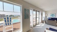 This sitting area embodies a relaxed coastal aesthetic, thoughtfully designed to maximize natural light and outdoor connectivity. The space is finished with neutral, light-toned walls and plush carpeting underfoot, creating a calm, inviting atmosphere throughout. Large picture windows and glass doors frame sweeping views of a surrounding deck, pool area, and open water beyond, seamlessly blending the interior with the outdoors.

The seating arrangement centers on wicker furniture upholstered in deep navy blue, including sofas, armchairs, and ottomans, providing generous and comfortable lounging options. Coastal-themed artwork adorns the walls, featuring herons, sea turtles, and waterscape paintings in aqua and blue tones that reinforce the nautical character of the space. A tall potted fiddle-leaf fig plant adds a natural, organic element to the room.

Recessed lighting and a wall-mounted mini-split system ensure year-round comfort. A distinctive interior window niche on one wall adds architectural interest, framing a vase as a decorative focal point. Glass panel doors open directly to the deck, allowing easy outdoor access and filling the room with abundant natural light throughout the day.