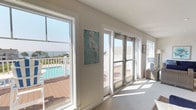 This sitting area embodies a relaxed coastal aesthetic, thoughtfully designed to maximize natural light and outdoor connectivity. The space is finished with neutral, light-toned walls and plush carpeting underfoot, creating a calm, inviting atmosphere throughout. Large picture windows and glass doors frame sweeping views of a surrounding deck, pool area, and open water beyond, seamlessly blending the interior with the outdoors.

The seating arrangement centers on wicker furniture upholstered in deep navy blue, including sofas, armchairs, and ottomans, providing generous and comfortable lounging options. Coastal-themed artwork adorns the walls, featuring herons, sea turtles, and waterscape paintings in aqua and blue tones that reinforce the nautical character of the space. A tall potted fiddle-leaf fig plant adds a natural, organic element to the room.

Recessed lighting and a wall-mounted mini-split system ensure year-round comfort. A distinctive interior window niche on one wall adds architectural interest, framing a vase as a decorative focal point. Glass panel doors open directly to the deck, allowing easy outdoor access and filling the room with abundant natural light throughout the day.
