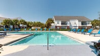 Located at 600 Crowne Commerce Ct in Ormond Beach, Florida, this outdoor amenity space offers a resort-style pool experience designed for relaxation and recreation under the Florida sun. The centerpiece is a large, crystal-clear swimming pool with gently graduated depths, accessible via wide entry steps and equipped with handrail entries for convenience. The expansive pool deck surrounds the water on multiple sides and is furnished with numerous blue lounge chairs and side tables, providing ample space for sunbathing and leisure. Towering palm trees and lush landscaping with manicured hedges frame the perimeter, lending a tropical ambiance to the setting. A dedicated outdoor grilling and dining area is positioned along one side of the deck, featuring stone-clad built-in grills alongside additional seating under a large patio umbrella — ideal for outdoor gatherings. The space is enclosed by a classic black iron fence, ensuring privacy while preserving open views of the surrounding greenery and adjacent buildings. The architecture of the neighboring structures features white siding and brick accents with pitched rooflines, complementing the well-maintained community aesthetic. Under clear blue skies, this outdoor space embodies the relaxed, sun-soaked lifestyle characteristic of Ormond Beach.