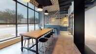 Located at 530 Meeting St in Charleston's vibrant Upper Peninsula neighborhood, this single-floor commercial space is a thoughtfully designed coworking and collaborative environment. The interior is defined by bold architectural character, most notably an eye-catching diagonal wood lattice ceiling installation that adds warmth and visual depth against exposed dark structural elements and ductwork above.

Floor-to-ceiling windows wrap around key areas of the space, flooding the interior with abundant natural light and offering open views of the surrounding Charleston streetscape. The polished stone-tile flooring runs throughout, lending a clean, professional feel underfoot.

Seating options are varied and purposeful: a large communal high-top table with bar stools serves as a central hub for collaborative work, while a dedicated meeting area features a solid butcher-block table surrounded by ergonomic chairs, ideal for focused group sessions. A lounge corner with teal upholstered chairs and minimalist side tables provides a relaxed setting for informal conversations.

Frosted glass partitions with black framing divide sections of the space while maintaining an open, airy atmosphere. Teal accent walls punctuate the interior with color, complementing the natural wood tones. Pendant lighting and recessed fixtures provide layered illumination throughout. This space blends contemporary design with functional versatility, making it well-suited for professionals, teams, and creative collaborators alike.