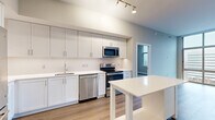 Located at 3400 South Clark Street in Arlington, VA, this well-appointed 2-bedroom, 2-bathroom residence offers a thoughtfully designed single-level layout. The open-concept kitchen and dining area serves as the heart of the home, showcasing crisp white shaker-style cabinetry extending to the ceiling, white quartz countertops, a subway tile backsplash, and a full suite of stainless steel appliances including a refrigerator, range, dishwasher, and built-in microwave. A freestanding white kitchen island provides additional prep space and storage. Warm-toned hardwood-style flooring flows throughout the main living areas, complementing the neutral palette. The spacious living room is a standout feature, anchored by dramatic floor-to-ceiling windows that frame sweeping urban views of the Arlington skyline, flooding the space with natural light. The generously sized primary bedroom includes a double-door closet and connects to the hallway. Both bathrooms are finished with large-format tile flooring and white vanities; one features a tiled tub-shower combination. Additional highlights include an in-unit laundry area, multiple closets for ample storage, and a welcoming entryway with a distinctive blue front door.