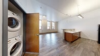 Located at 200 Cowan St in Pittsburgh's South Side neighborhood, this one-bedroom, one-bathroom unit offers a well-designed open-concept layout that maximizes every square foot of living space. The main living and dining area features hardwood-style plank flooring that flows seamlessly throughout, soaring ceilings, and a row of tall windows with red-trimmed frames that flood the space with natural light. Track lighting and modern pendant fixtures provide additional illumination throughout. The kitchen is equipped with warm wood cabinetry, white countertops, a tiled mosaic backsplash, and black appliances including a refrigerator, electric range, and microwave. A peninsula with a sink separates the kitchen from the dining area, creating a functional and social layout. The bedroom is a comfortable, well-proportioned retreat featuring large windows with roller shades, hardwood-style flooring, and clean white walls. The full bathroom includes a warm wood vanity with a white countertop, a tub-shower combination, tile flooring in a checkered pattern, and crisp white walls. A dedicated closet and in-unit washer and dryer connections add to the unit's practicality. The overall aesthetic is clean and contemporary, making this a functional and well-lit urban residence.