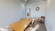 This single-floor professional space is thoughtfully designed to support focused meetings and collaborative work. The centerpiece of the space is a private conference room finished with classic crown molding and soft blue-gray walls that create a calm, productive atmosphere. A warm wooden conference table is surrounded by upholstered task chairs, with an additional accent chair positioned along the wall, offering flexible seating for various group sizes. An elegant chandelier overhead lends a refined touch, complementing the room's polished aesthetic. A decorative wall clock adds both a functional and stylistic element to the space. The conference room connects through a glass-paneled door to an adjacent room that appears to serve as a kitchenette or break area, featuring warm wood cabinetry and pendant lighting — providing added convenience for extended meetings or working sessions. Neutral-toned carpeting throughout contributes to the room's quiet, professional feel. The overall design balances practicality with a sophisticated, welcoming ambiance, making it well-suited for private client meetings, team discussions, or collaborative work sessions.