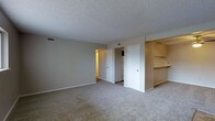 This single-floor residential space offers a straightforward and functional layout designed for comfortable everyday living. The main living area is finished with wall-to-wall carpeting in a neutral tone, providing a warm and cohesive foundation throughout the room. Walls are painted in a soft, light gray that lends a clean and understated aesthetic to the space.

A textured popcorn ceiling adds a classic residential character overhead, while the standard ceiling height creates an intimate and cozy atmosphere. Visible HVAC vents integrated into the ceiling ensure consistent climate control throughout.

Multiple doorways branch off from the main living area, offering connectivity to additional rooms. One passageway leads to an adjoining room with warmer-toned lighting, suggesting a hallway or secondary living space, while a white paneled door provides access to a closet or utility area.

A standout feature of this space is the built-in shelving nook positioned along one wall, offering dedicated storage or display space. The shelving is finished in a light wood tone, complementing the neutral color palette of the room. White baseboards frame the perimeter of the space, adding a crisp, finished detail to the overall design. This space presents a clean, move-in-ready environment well-suited for a variety of furnishing arrangements.