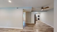 This single-floor space presents a thoughtfully updated interior with a cohesive, modern aesthetic. Light hardwood-style flooring runs throughout the main living areas, complementing the soft gray and white wall tones that give the space a bright, airy feel. Textured ceilings and recessed lighting are consistent throughout, with ceiling fans adding both function and style to select areas.

The open-concept layout, visible in the overhead view, connects the living area, kitchen, and hallway corridor efficiently. The kitchen is well-equipped with stainless steel appliances — including an electric range, over-the-range microwave, and refrigerator — set against crisp white cabinetry and gray stone-style countertops that also appear in the bathroom vanity, creating a unified design language.

A dedicated laundry area features a stacked washer and dryer unit, offering convenient in-unit functionality. The bedroom area includes a ceiling fan, a closet with shelving, and direct access to the entry door. The bathroom is cleanly finished with white tile flooring, a tub-shower combo, white cabinetry, and matching countertops. Overall, the space delivers a clean, move-in-ready environment with consistent, modern finishes.