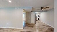 This single-floor space presents a thoughtfully updated interior with a cohesive, modern aesthetic. Light hardwood-style flooring runs throughout the main living areas, complementing the soft gray and white wall tones that give the space a bright, airy feel. Textured ceilings and recessed lighting are consistent throughout, with ceiling fans adding both function and style to select areas.

The open-concept layout, visible in the overhead view, connects the living area, kitchen, and hallway corridor efficiently. The kitchen is well-equipped with stainless steel appliances — including an electric range, over-the-range microwave, and refrigerator — set against crisp white cabinetry and gray stone-style countertops that also appear in the bathroom vanity, creating a unified design language.

A dedicated laundry area features a stacked washer and dryer unit, offering convenient in-unit functionality. The bedroom area includes a ceiling fan, a closet with shelving, and direct access to the entry door. The bathroom is cleanly finished with white tile flooring, a tub-shower combo, white cabinetry, and matching countertops. Overall, the space delivers a clean, move-in-ready environment with consistent, modern finishes.