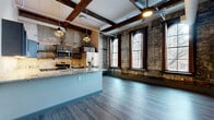 Located at 1273 W Ninth St in Cleveland's vibrant Ohio City neighborhood, this thoughtfully designed unit blends historic industrial character with modern finishes. The open-concept living and kitchen area serves as the heart of the space, where original exposed brick walls, reclaimed wood ceiling beams, and striking arched windows with dark wood frames create a dramatic, loft-like atmosphere. Natural light pours in through the tall arched windows, illuminating the smooth hardwood-style flooring throughout. The kitchen features a central island with granite countertops, a built-in sink, pendant lighting, stainless steel appliances, and custom cabinetry with a wine rack detail. The bedroom offers a comfortable footprint with direct access to one of the five bathrooms. Each bathroom is finished with granite-topped dark wood vanities, framed mirrors, and hardwood-style flooring, maintaining a consistent design language throughout. A walk-in closet with exposed brick and built-in shelving adds a unique industrial touch. Two hallways connect the various spaces efficiently, with pendant globe lighting and visible ductwork reinforcing the building's converted industrial heritage. The overall ambiance is a refined blend of raw, historic texture and contemporary urban living.