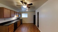 This single-floor space features an open-concept layout that seamlessly connects the kitchen and living area, creating a functional and cohesive environment. The kitchen is outfitted with rich, dark wood cabinetry that provides ample storage, complemented by a double-basin sink, black appliances including a range, dishwasher, and refrigerator, and dark countertops that tie the design together. A window above the sink area allows natural light to filter into the kitchen.

The living area flows directly from the kitchen, offering an open and versatile space. Warm, hardwood-style flooring extends throughout the entire area, adding a sense of continuity and warmth to the interior. A ceiling fan with an integrated light fixture is centrally positioned, providing both air circulation and overhead lighting to the combined space.

A doorway with a white-trimmed frame on the far wall leads to an adjacent room or closet area with visible shelving, adding convenient storage options. The neutral-toned walls throughout keep the space feeling bright and adaptable, offering a clean backdrop suitable for various décor styles.