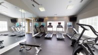 This fitness center offers a comprehensive workout environment designed to support a wide range of exercise routines. The space is organized across interconnected areas, each serving a distinct fitness purpose. The main cardio zone features a row of treadmills equipped with individual monitors, complemented by an elliptical and a stationary bike, all positioned near large windows that allow ample natural light and outdoor views. Vaulted ceilings with ceiling fans enhance airflow and contribute to an open, airy atmosphere. Full-length mirrors line the walls, visually expanding the space and allowing for form checks during workouts. A dedicated strength training area houses a multi-functional cable machine alongside a weight bench and a well-stocked dumbbell rack. The free weight section is neatly organized and easily accessible. A vibrant yellow-walled flex room provides space for stretching, yoga, and functional training, equipped with resistance bands, exercise mats, and balance tools. Dark rubber flooring throughout provides durability and traction. The neutral warm-gray color palette, white trim, and tasteful wall art create a clean, motivating environment suited for daily fitness routines.