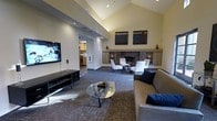 This single-floor space makes an immediate impression with its soaring vaulted ceiling, which lends a sense of grandeur and openness to the main living area. Recessed lighting and strategically placed accent lights illuminate the space, complementing the warm, neutral tones of the walls and carpeted floors.

The living area is thoughtfully arranged with a large, comfortable sofa adorned with navy accent pillows, centered around a sleek glass coffee table. A wall-mounted flat-screen television anchors one side of the room, positioned above a low-profile media console. On the opposite end, a built-in stone fireplace serves as a striking focal point, flanked by built-in shelving and cabinetry in a muted gray finish. A pair of white accent chairs face the fireplace, creating an inviting conversation nook. Two framed art pieces are displayed above the mantle, adding a refined decorative touch.

The open layout connects seamlessly to an adjoining kitchen, visible through a wide entryway. The kitchen features warm wood cabinetry, offering a welcoming contrast to the cooler tones of the main living space. Throughout, the design balances comfort and sophistication, with a cohesive neutral palette and contemporary furnishings that create a polished yet approachable atmosphere.