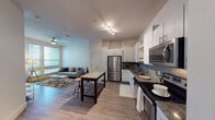 This thoughtfully designed single-floor residence offers a seamless open-concept layout that connects the kitchen and living area, creating a spacious and functional flow throughout. The kitchen is a standout feature, showcasing crisp white cabinetry that extends to the ceiling, a striking grey subway tile backsplash, dark granite countertops, and a full suite of stainless steel appliances including a refrigerator, range, and microwave. A freestanding granite-topped island with bar seating adds both prep space and a casual dining option. Track lighting overhead enhances the kitchen's modern aesthetic. Warm wood-look flooring runs throughout the main living areas, anchoring the open living room, which benefits from generous natural light streaming through large windows and a glass-paneled door. A ceiling fan adds comfort to the airy living space. The bathroom is well-equipped with a granite-topped vanity, a large mirror, wood-look flooring, and a glass-enclosed walk-in shower, directly adjacent to a spacious walk-in closet with built-in shelving. The bedroom features plush carpet flooring and bright windows that invite natural light. Clean, neutral wall tones throughout give the space a calm and modern ambiance.