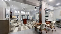 Located at 2800 Pontiac Dr in Aurora, IL, this thoughtfully designed single-floor amenity space offers a refined and functional environment for socializing, working, and relaxing. The open-concept lounge area commands attention with soaring ceilings, floor-to-ceiling windows, and an abundance of natural light that fills the room. Geometric patterned carpet tiles in neutral gray and brown tones anchor the space, complementing the contemporary furnishings that include sleek seating arrangements, a high-top communal bar table with stylish swivel chairs, and curated built-in wooden shelving units displaying decorative accents. Sculptural multi-bulb chandeliers provide warm, ambient lighting throughout. The kitchen is equally impressive, featuring a large central island with a striking white quartz countertop, a dark navy ceiling, frosted glass upper cabinetry, rich walnut lower cabinets, stainless steel appliances, and a dedicated coffee bar with a garden-view window. A built-in workstation area with dual computer setups offers a quiet productivity zone, while additional seating nooks provide flexible gathering spaces. The overall ambiance is modern and sophisticated, blending warm wood tones with cool grays for a polished, welcoming atmosphere.