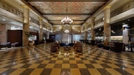 This magnificent hotel lobby is a masterclass in Beaux-Arts grandeur, offering a sweeping, voluminous space defined by soaring ceilings adorned with elaborately painted coffered panels featuring classical motifs in rich golds, reds, and blues. Suspended crystal chandeliers cast a warm, refined glow across the entire hall, complementing the ornate decorative plasterwork that frames the ceiling's perimeter.

Towering marble columns with veined golden and sienna tones line the space, supporting an upper-level mezzanine gallery framed by elegant ironwork balustrades. The signature checkerboard terrazzo floor in warm amber and deep brown tones anchors the space with timeless sophistication, stretching across every area of the lobby.

The central seating area features plush leather armchairs and curved sofas arranged around low tables, creating an inviting atmosphere for guests. Along the perimeter, intimate seating vignettes are appointed with console tables, decorative mirrors, and floral arrangements, adding warmth to the classical architecture. A sleek modern reception desk provides a subtle contemporary contrast.

Additional areas reveal an atmospheric lounge with dark-paneled walls, a water feature with river stones, and sculptural pendant installations. Throughout, the interplay of historic architectural detail and refined contemporary furnishings creates an atmosphere of understated luxury and timeless elegance.